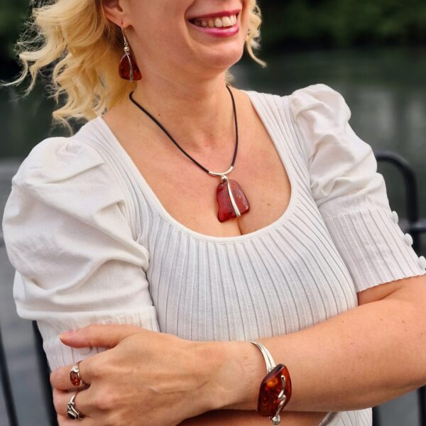 A person stands confidently in a white blouse, showcasing the COGNAC AMBER AND SILVER TWIST SET—necklace, earrings, ring, and bracelet. With arms crossed against a blurred outdoor backdrop, the amber accents shimmer subtly.