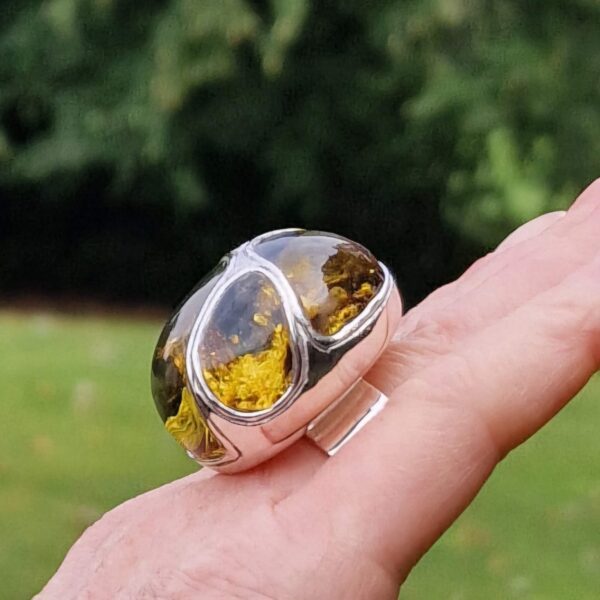 The product "A Legendary Masterpiece of Unrivaled Beauty – An Ode to Earth’s Ancient Secrets" is a large silver ring with a translucent amber stone, showcased on an outstretched hand with a blurred green backdrop.